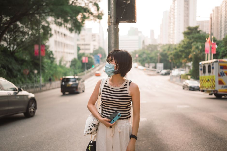 Woman wearing a face mask standing on a sunlit city street, surrounded by vehicles and buildings.